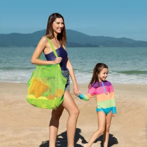 Mulher de maiô azul caminhando na praia de mãos dadas com a filha e carregando a bolsa de brinquedos Tobee verde