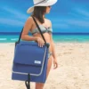 Mulher na praia carregando a espreguiçadeira azul Tobee fechada no ombro como uma bolsa prática com alça preta.