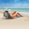 Mulher de chapéu deitada de costas na espreguiçadeira de praia cinza Tobee, posicionada sobre a areia em frente ao mar azul. O bolso traseiro com zíper e a alça de transporte estão bem visíveis.