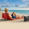 Homem de chapéu relaxando na espreguiçadeira de praia vermelha articulada da marca Tobee na areia, de frente para o mar azul.