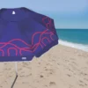 Detalhe em close do tecido do guarda-sol Campeche Tobee azul e rosa montado na areia com mar azul ao fundo, destacando a qualidade da lona e a estampa do ombrelone de praia.