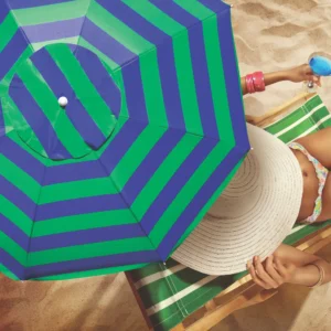 Mulher de chapéu deitada em cadeira de praia sob a sombra do Guarda Sol Aruba Tobee listrado azul marinho e verde segurando uma bebida.