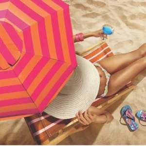 Visão aérea de modelo na espreguiçadeira sob a sombra do Guarda Sol Aruba Tobee listrado vermelho e laranja na areia.