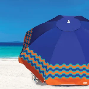 uarda-sol de praia azul com estampa de ondas laranja e azul-celeste da marca Tobee, aberto na areia branca em frente ao mar.