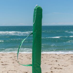 Um ombrelone grande portátil da marca Tobee, modelo Malibu Beach, fechado e armazenado dentro de sua capa de transporte de tecido verde com alça, em pé na areia da praia com o mar e o céu azul ao fundo.
