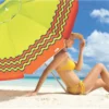 Mulher de biquíni amarelo e chapéu de palha sentada na areia da praia, abrigada sob a sombra de um guarda-sol verde e laranja Tobee.