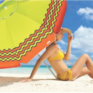 Mulher de biquíni amarelo e chapéu de palha sentada na areia da praia, abrigada sob a sombra de um guarda-sol verde e laranja Tobee.
